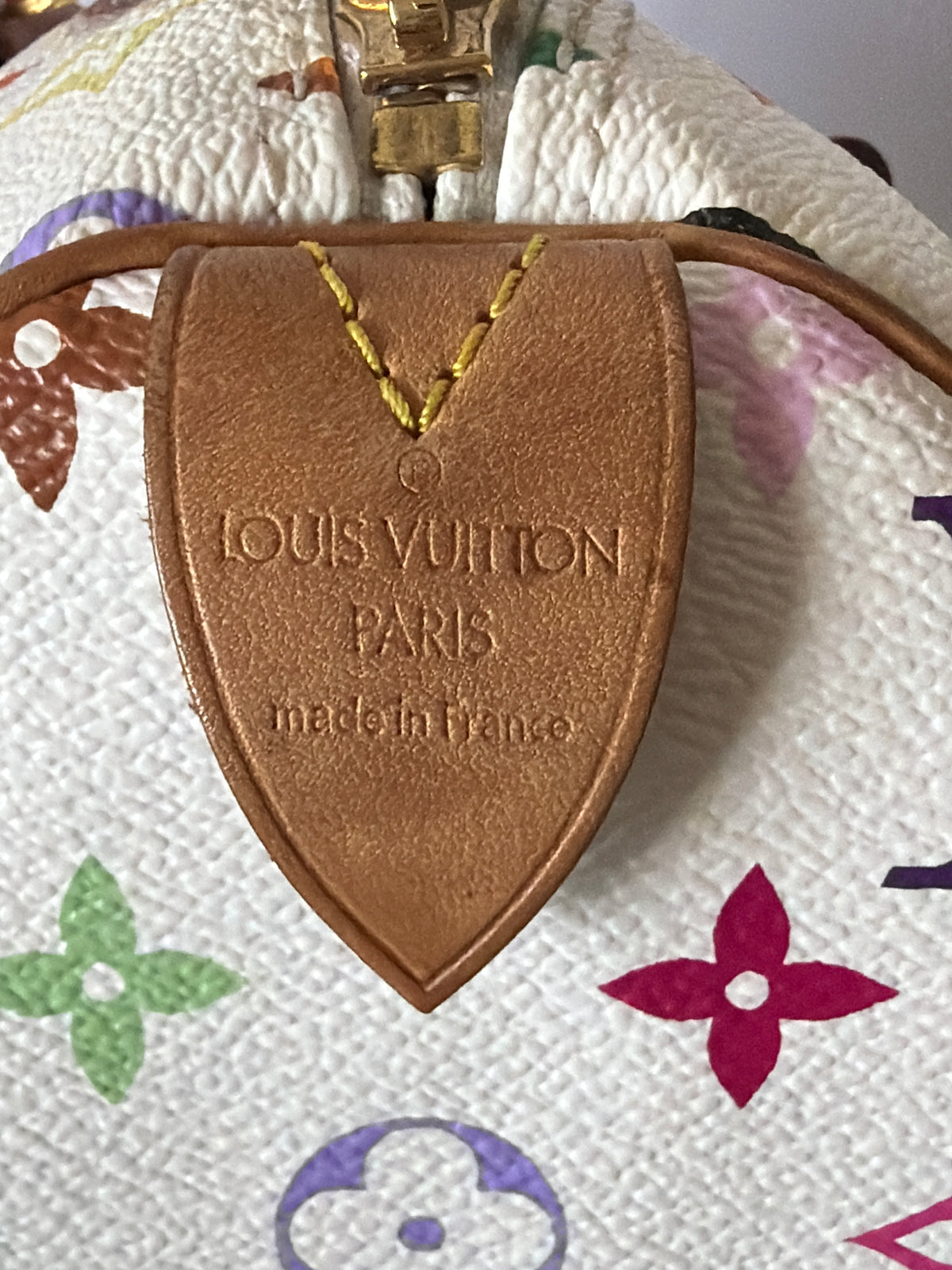 Close-up of a Louis Vuitton monogram bag showing a tan leather shield-shaped tag with 'LOUIS VUITTON PARIS made in France' embossed on it, attached to a gold-tone zipper pull on a white patterned canvas background.
