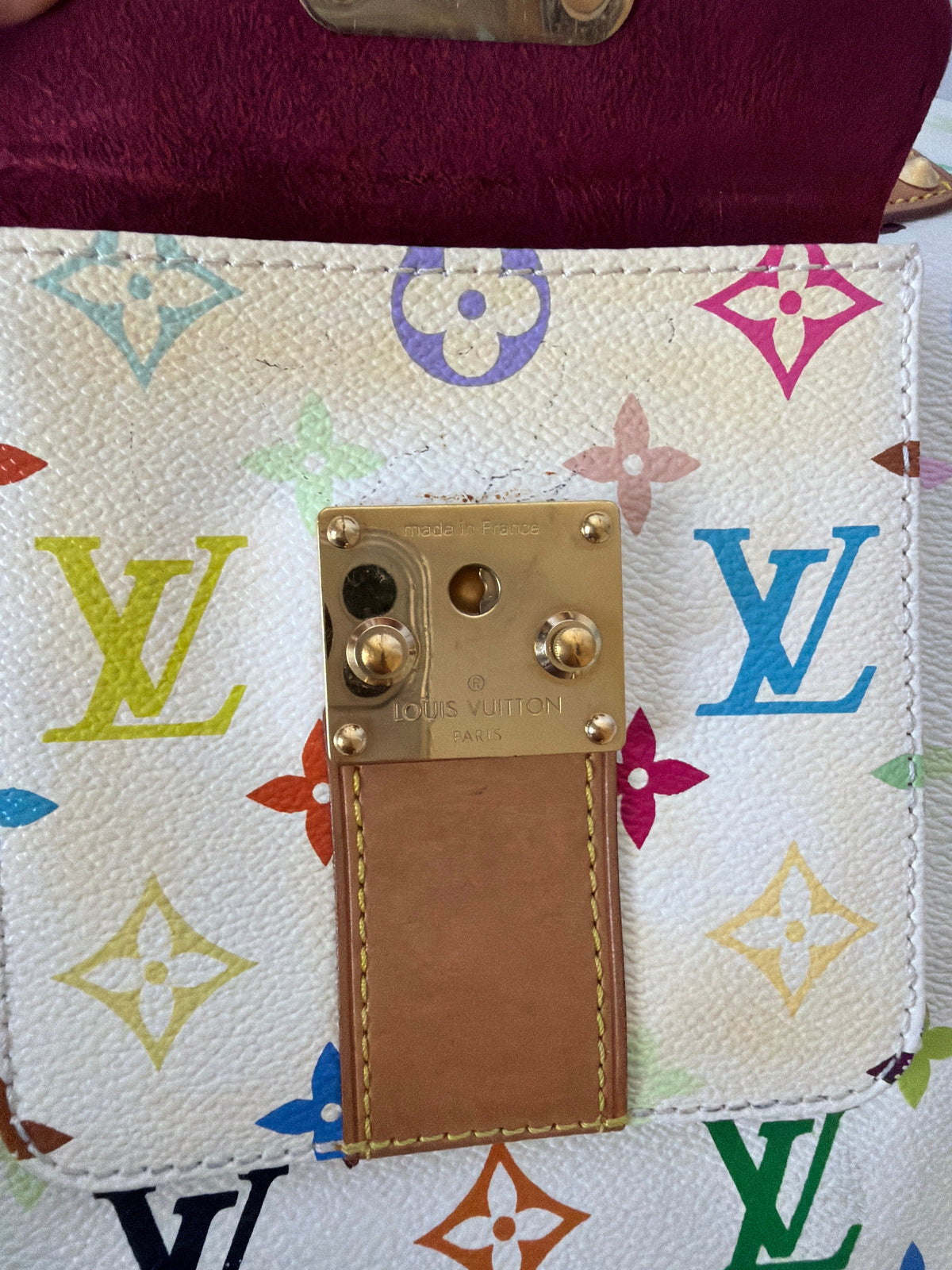 A close-up of a Louis Vuitton wallet or clutch showing the white monogram canvas with multicolored LV logos, a tan leather tab, and a gold-tone metal clasp. A burgundy leather section is visible on the left edge.