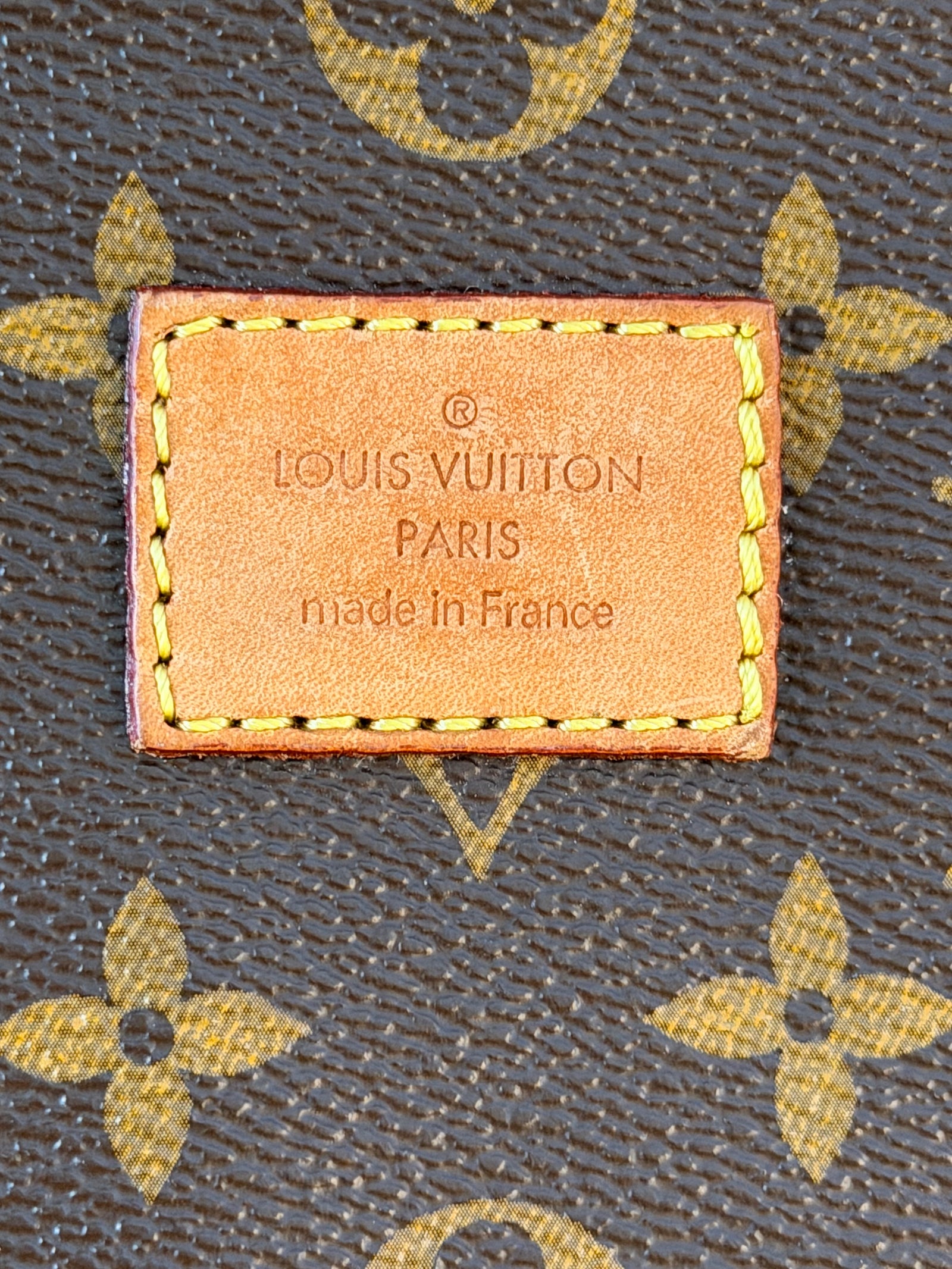Close-up of a Louis Vuitton monogram canvas surface with a stitched leather brand patch reading 'LOUIS VUITTON PARIS made in France'.