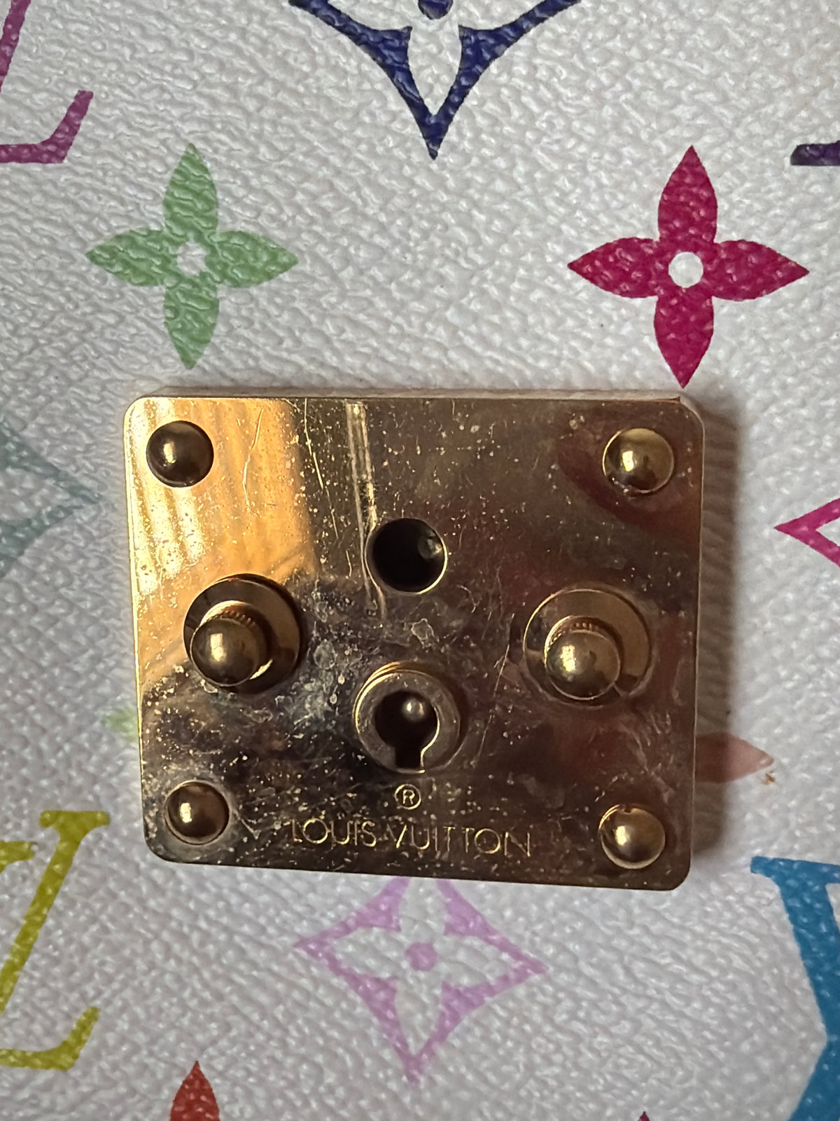 A square gold-toned metal lock component with six screw holes and the embossed Louis Vuitton logo, placed on a white Louis Vuitton monogram-patterned surface.