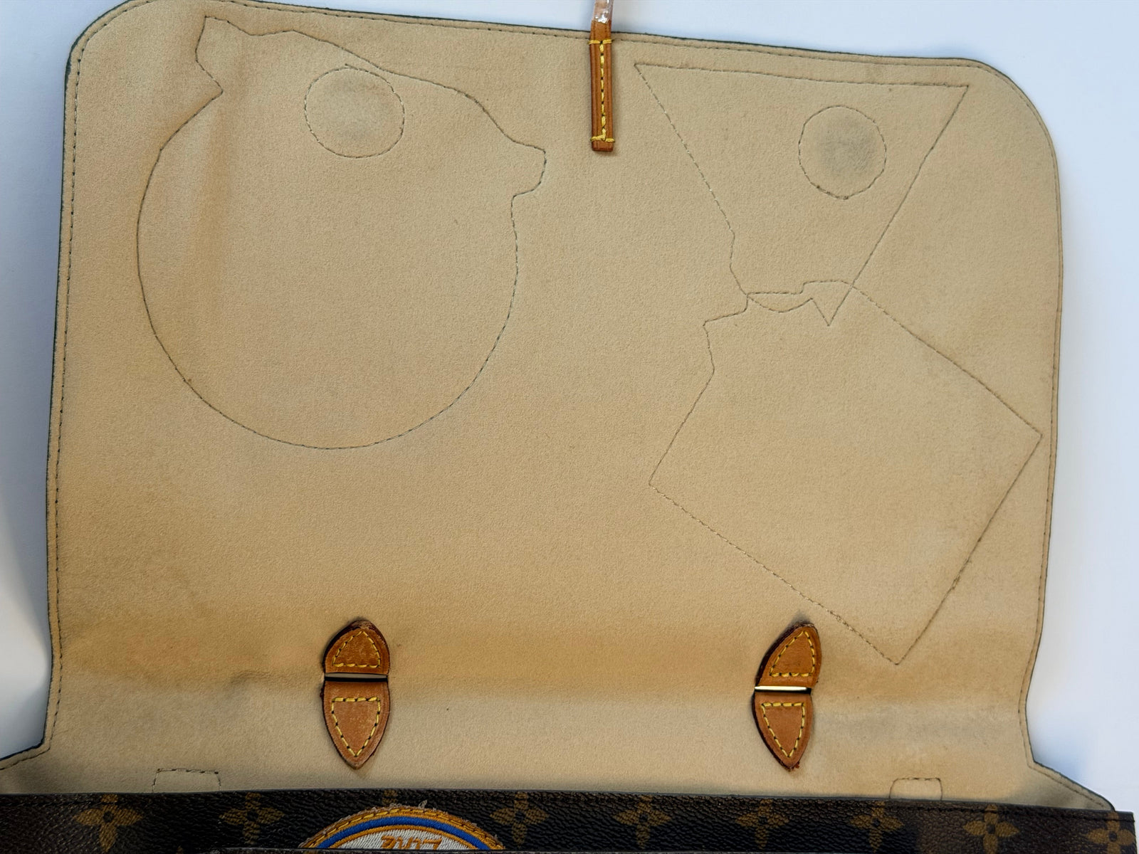 An open beige leather clutch or wallet showing the interior lining with stitched cutouts, two tan leather tab closures, and a partial view of a dark monogram patterned exterior on the right side.