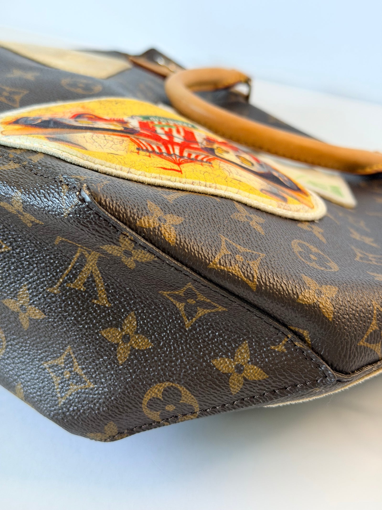 Close-up of a Louis Vuitton monogram canvas handbag with a colorful embroidered patch and a tan leather handle on a light background.