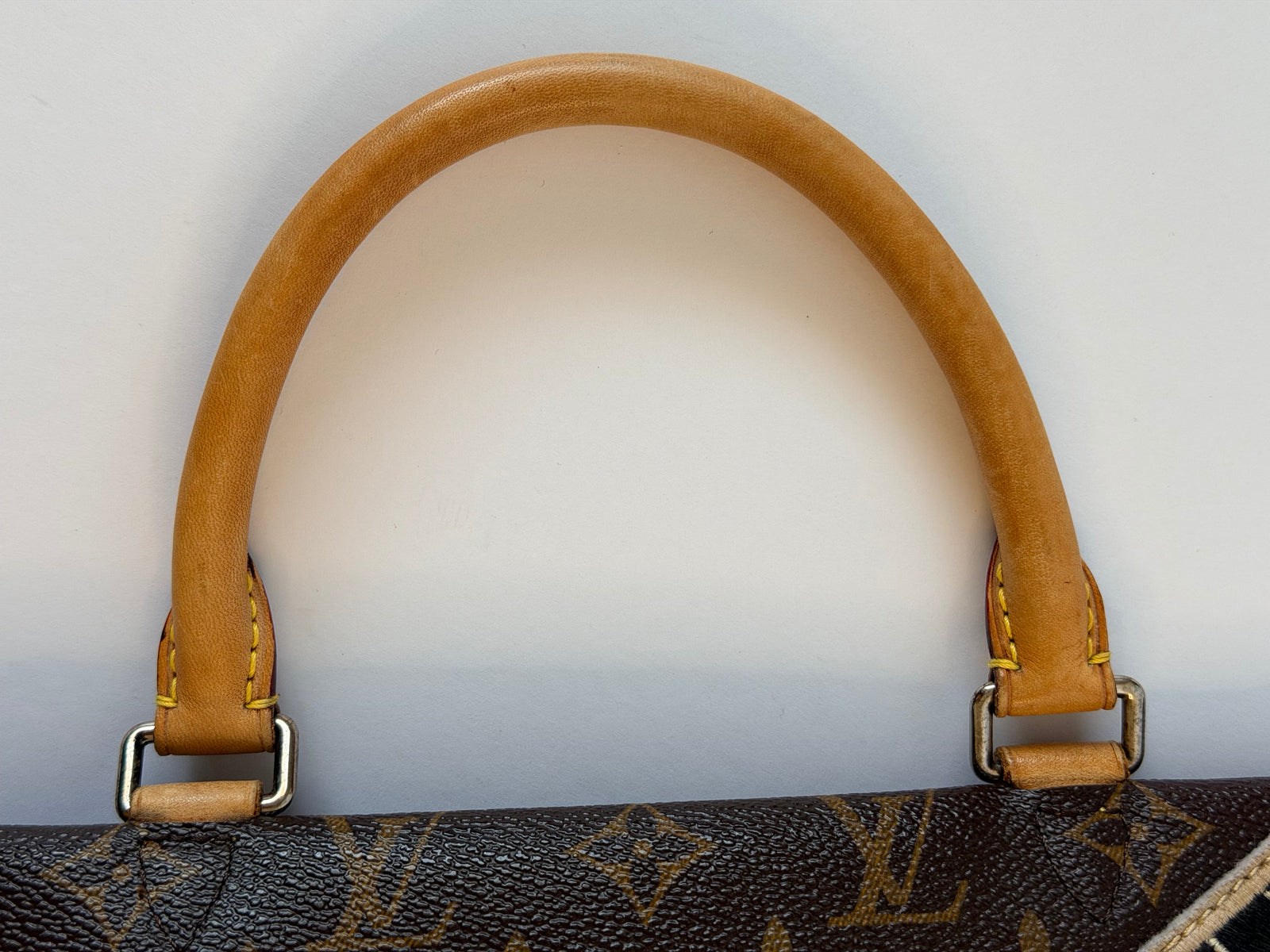 Close-up of a tan leather handle attached to a dark brown monogram canvas bag, showing metal hardware and yellow stitching on a light gray background.