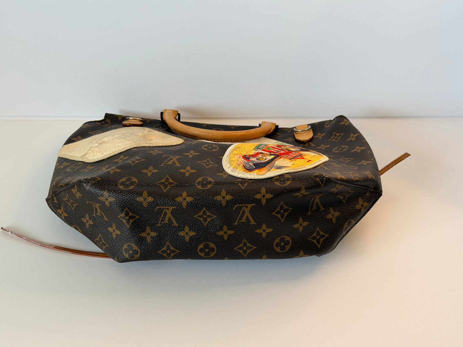 A Louis Vuitton monogram canvas duffel bag with tan leather handles, featuring a large beige patch and a circular colorful patch on the front, resting on a white surface against a white background.